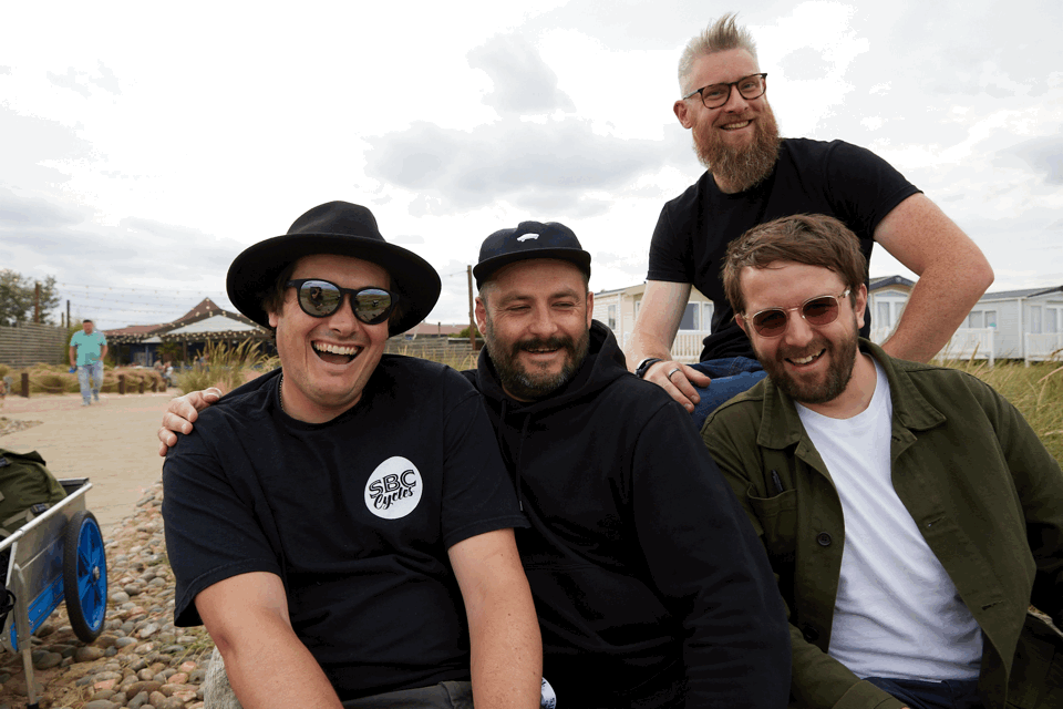 Four men smiling outdoors, sitting and standing among rocks and grass, with overcast skies in the background.
