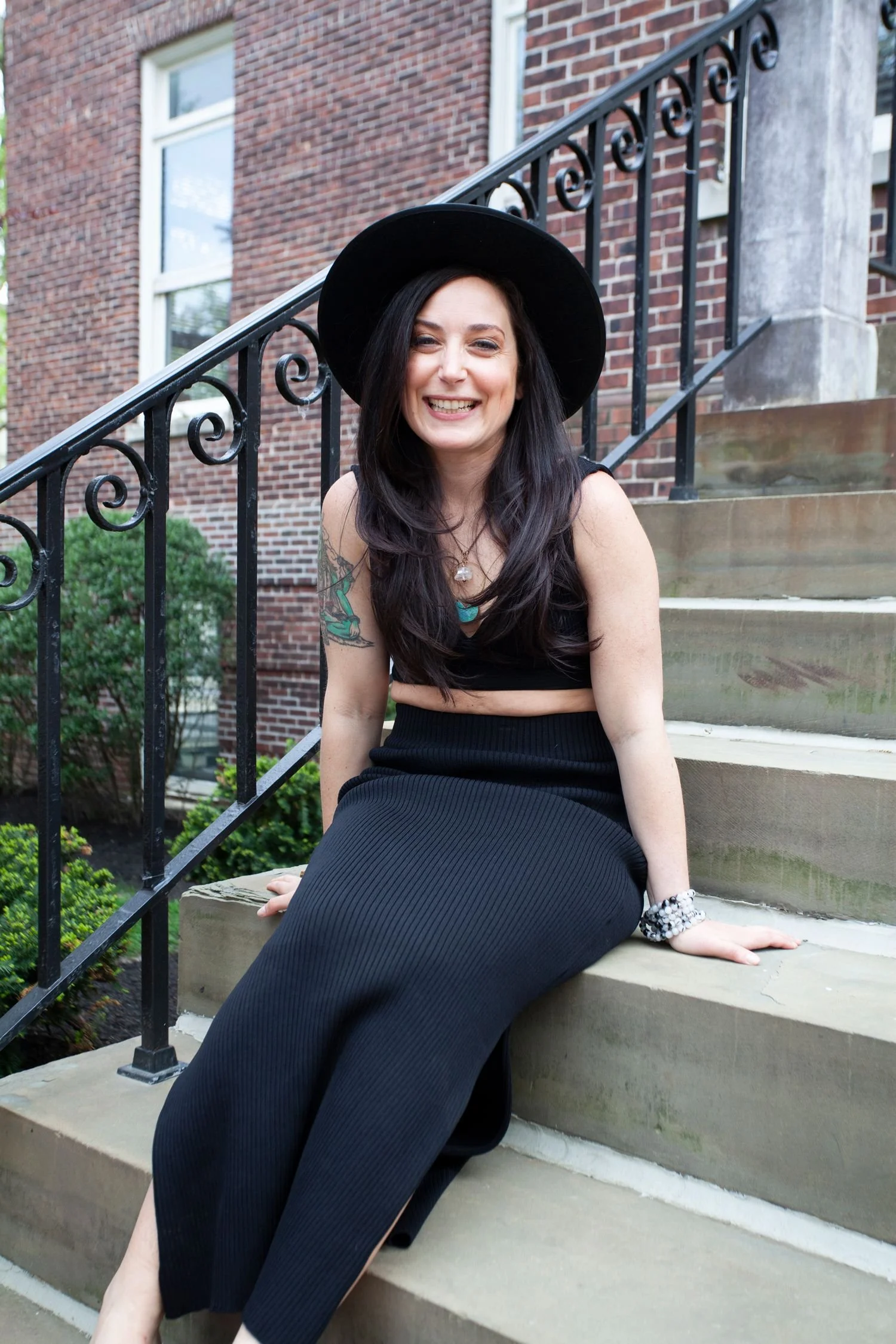 Woman sitting on stairs, wearing black hat and outfit, smiling.