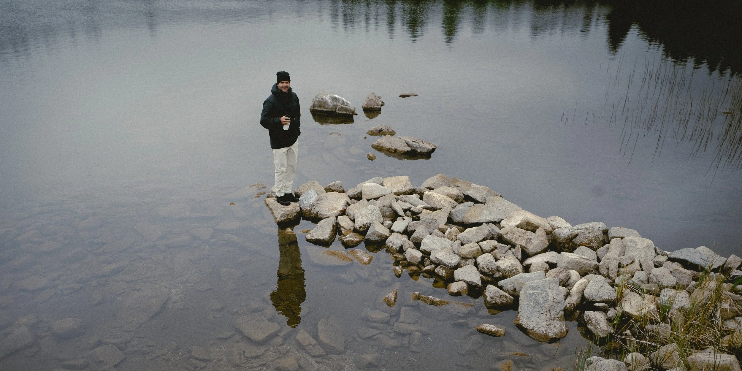 En mann står på en steinlagt sti i vannet, og holder et sort termokrus. Han er iført mørk jakke, hvite bukser og en svart lue. Man ser et refleksjon av ham i vannet.