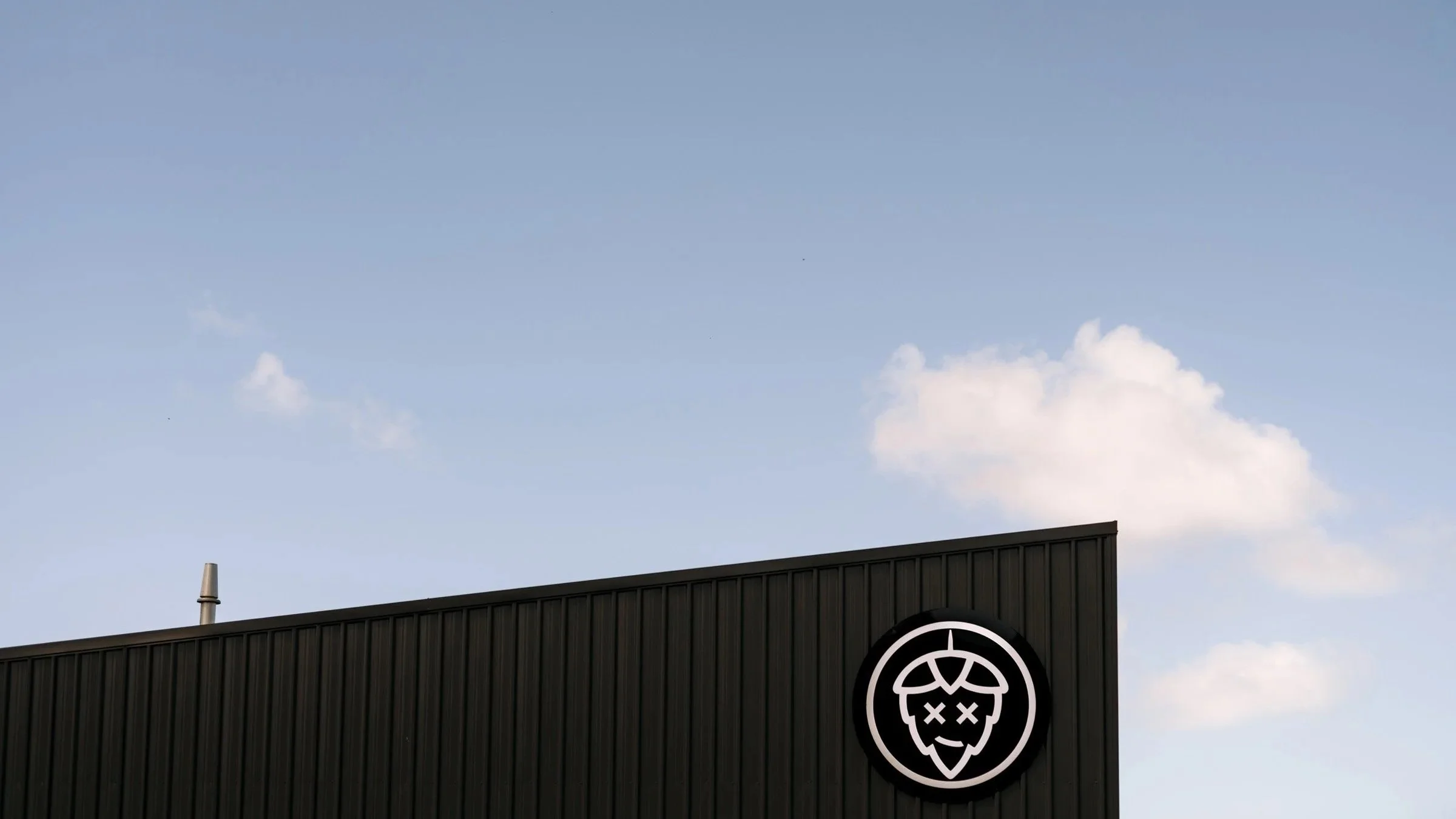 A black building with a circular logo of a stylized hop cone with a smiling face and crossed-out eyes, set against a blue sky with a few white clouds.