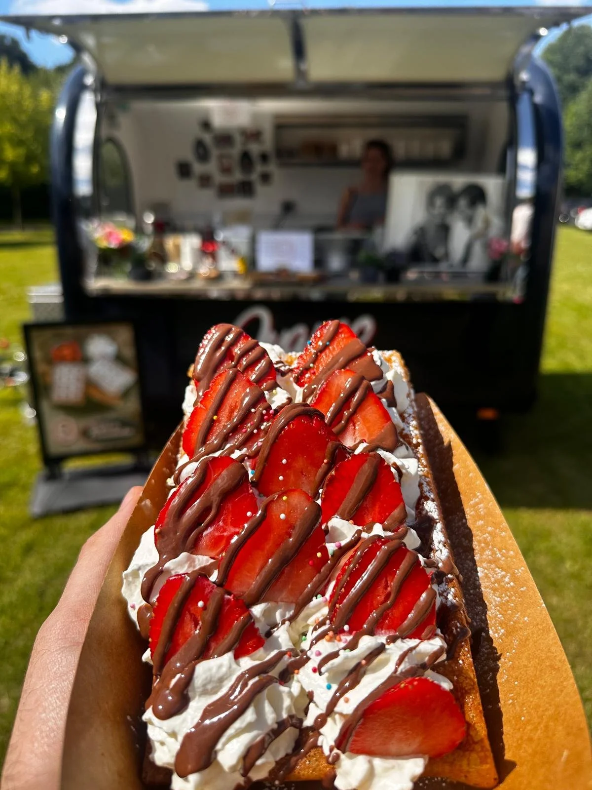 A person holding a strawberry-topped waffle with whipped cream and chocolate drizzle in front of a food truck. wafel met slagroom, aardbeien chcoladade saus en bloemsuiker.