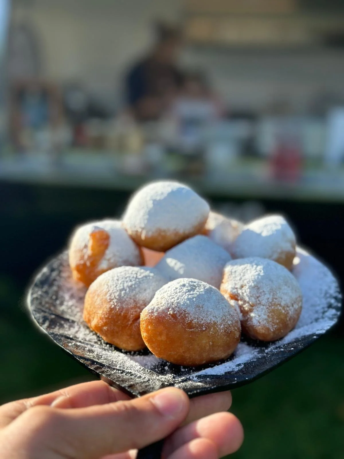 Fried dough balls topped with powdered sugar on a black paper plate held by a person's hand, with a blurred background of people sitting at a table. smoutebollen met bloemsuiker.