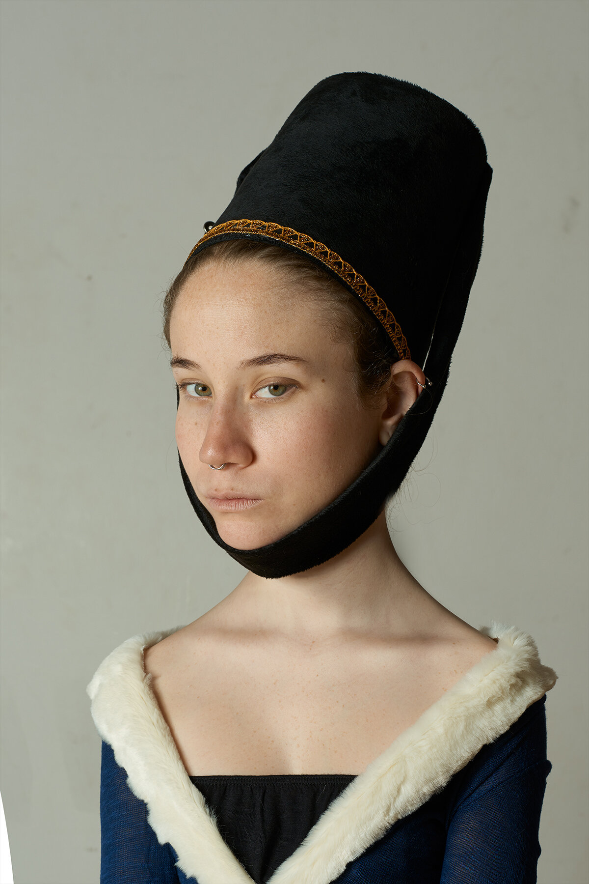 Retrato de una joven con sombrero grande y negro, vestimenta con detalles de piel blanca en el pecho, mirando a la cámara con expresión neutral.