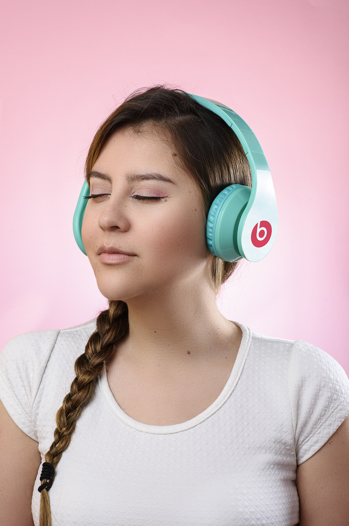 Joven mujer con cabello castaño y trenza, usando auriculares blancos y azul turquesa con logotipo de Beats, con ojos cerrados y expresión de relajación, fondo rosa.