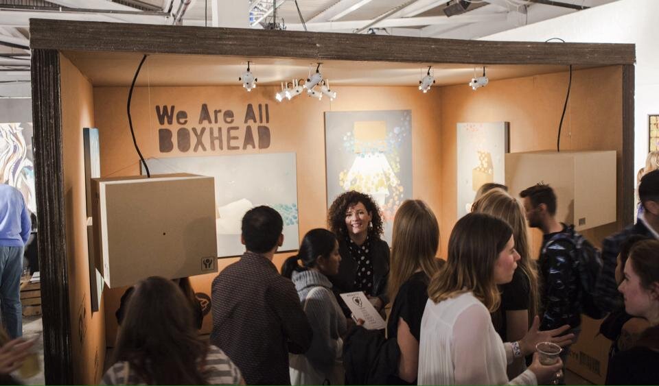 People gathering at an art exhibit booth with the sign 'We Are All Boxhead' in a gallery setting.