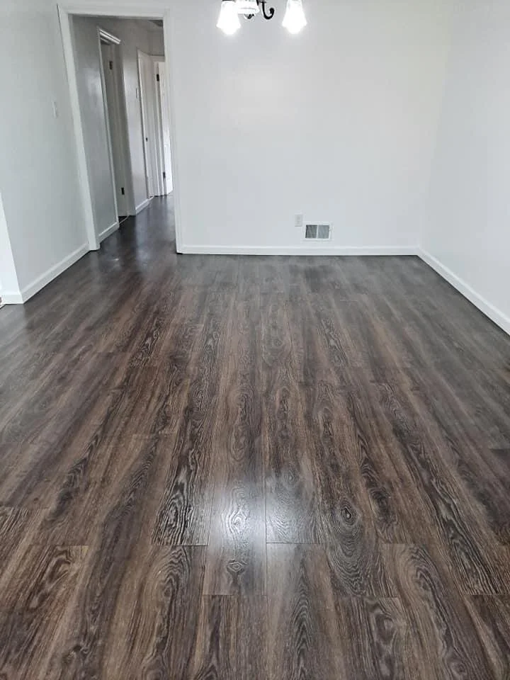 Empty room with white walls and dark wood flooring, hallway visible through an open doorway, ceiling light fixture with three bulbs.