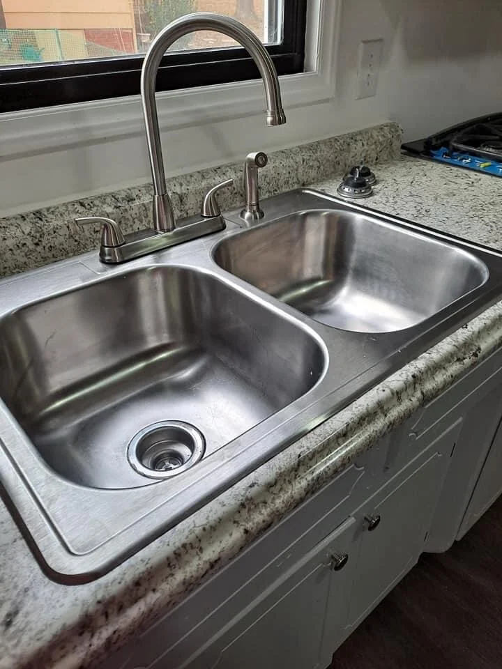 Stainless steel double kitchen sink with a high-arc faucet and a sprayer, set into a speckled granite countertop under a window.