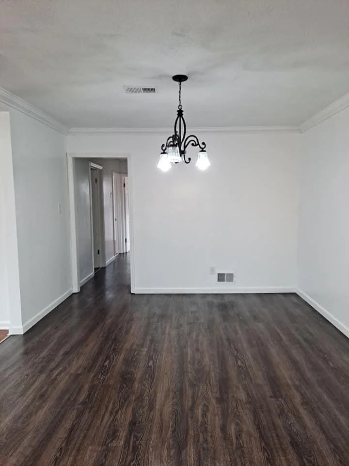 Empty dining room with white walls, dark wood flooring, and a black chandelier with three lights hanging from the ceiling.