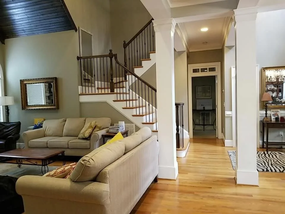 Living room with beige sofa, wooden stairs with dark railing, doorway leading to a hallway, hardwood floors, and various furniture and decorations.