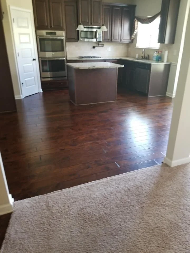 Kitchen with dark brown cabinets, stainless steel oven and microwave, granite countertop, a small island, and a window with curtains over the sink. Hardwood flooring in the kitchen area and carpet in the foreground.