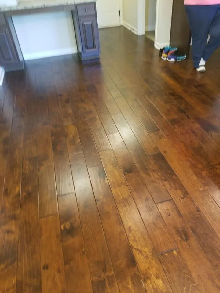 Photo of a wooden floor in a house, with a person standing in the corner near some shoes.