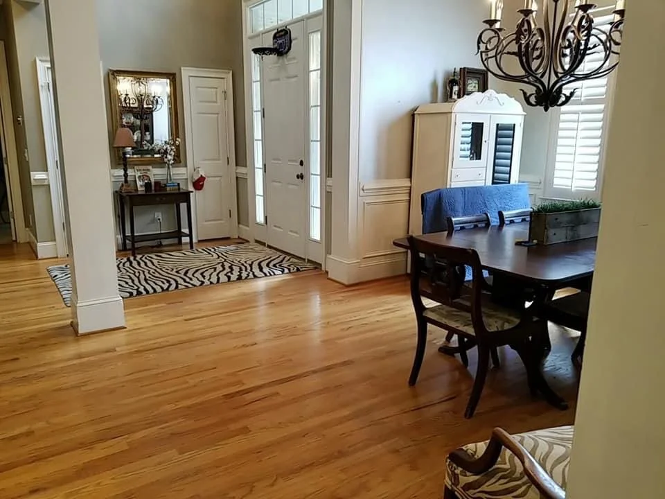 A dining room with hardwood floors, a dark wooden table with chairs, a white cabinet with glass doors, a chandelier, and a window with shutters.
