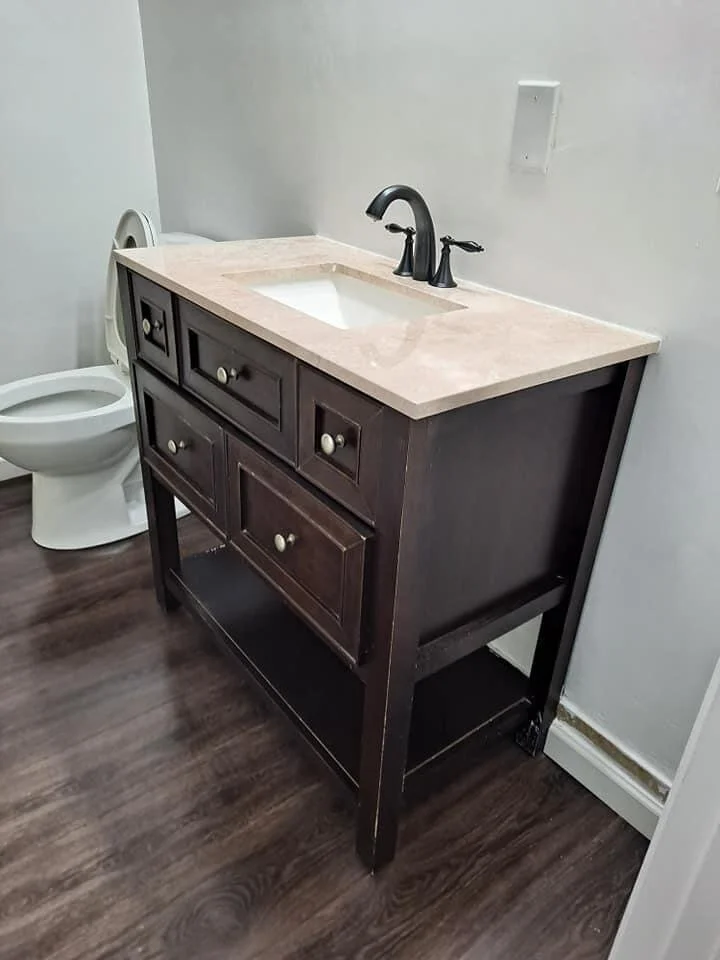 Bathroom with a dark wooden vanity with a beige marble countertop and an undermount sink, black faucet, and cabinet knobs. Toilet in the background and empty white wall with an electrical outlet.