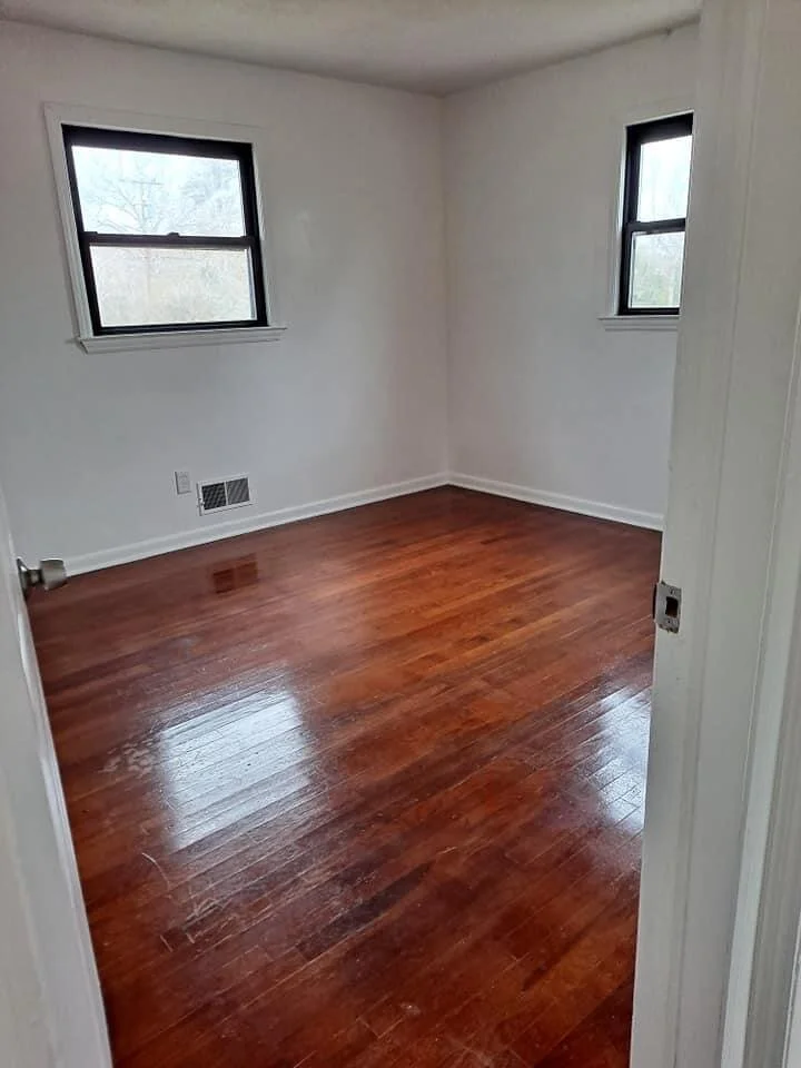 Empty room with white walls, two windows, and hardwood floors.
