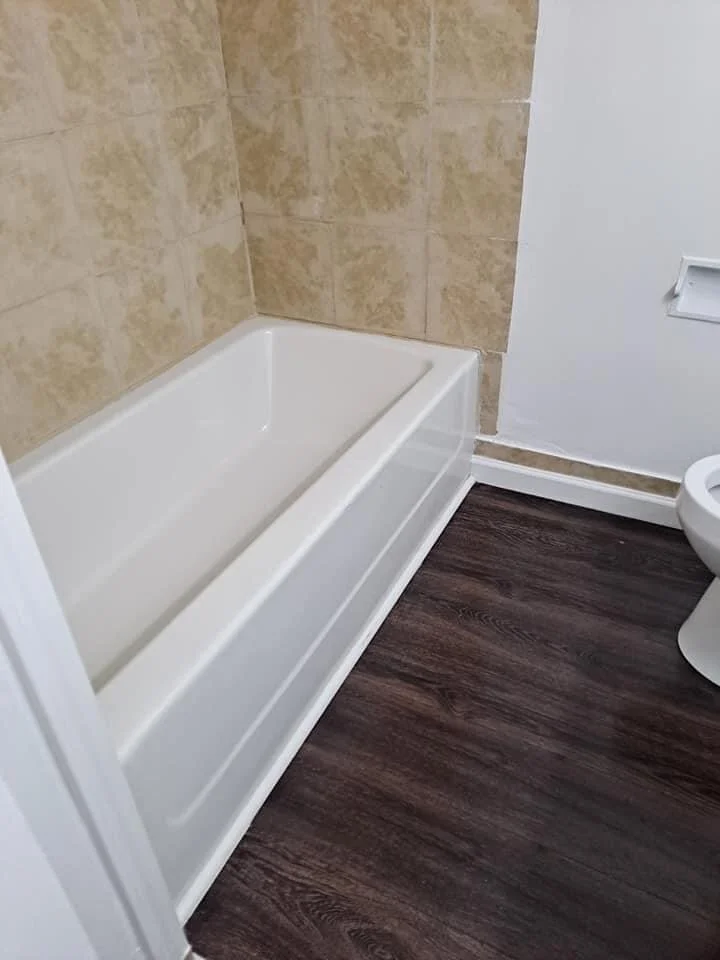 Bathroom with a white bathtub, beige tiled wall, white wall with a vent, and dark wood laminate flooring.