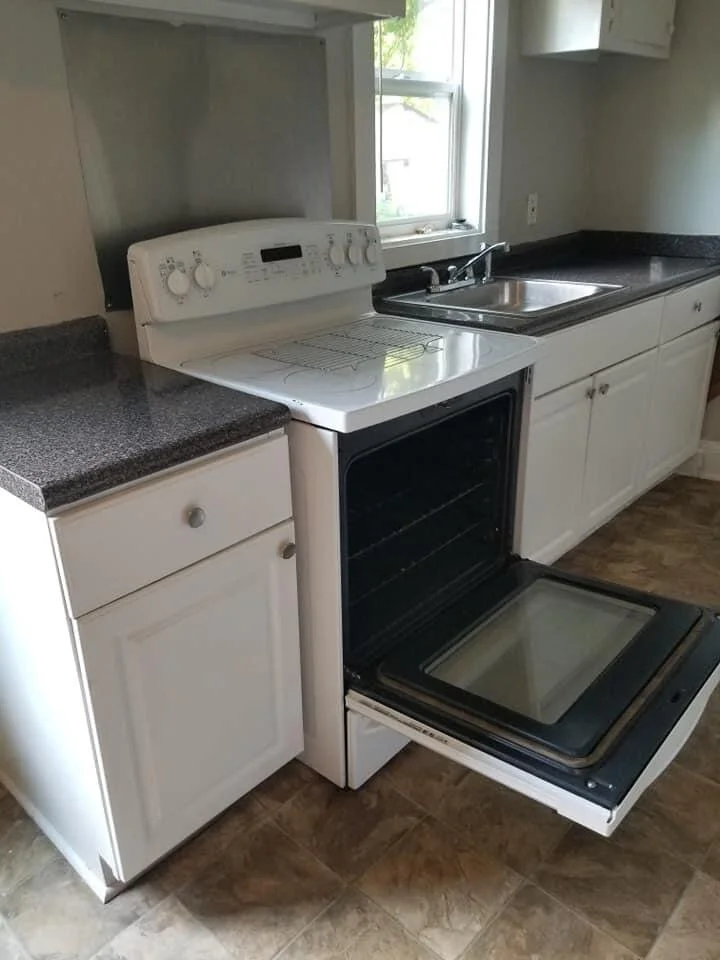 Kitchen with a white stove, black countertops, a stainless steel sink, and white cabinets, located near a window.