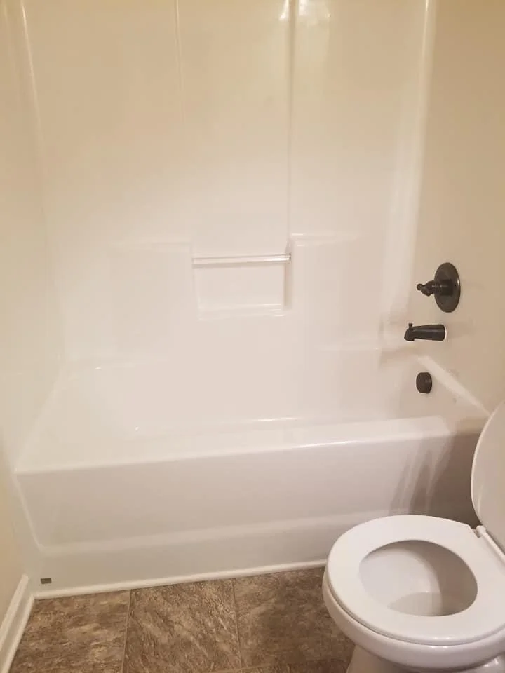 A bathroom shower and bathtub with a toilet in front, brown tiled floor, beige walls, and black fixtures.
