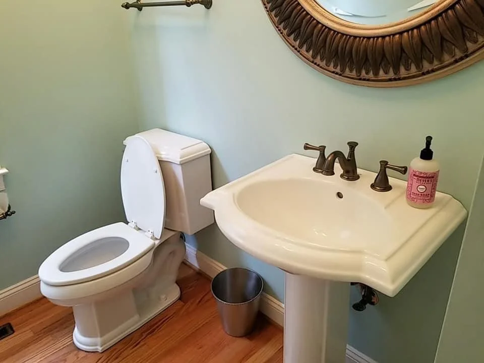 A small bathroom with a white toilet, a white pedestal sink with bronze fixtures, a pink hand soap bottle, a small metal trash can, a wooden framed mirror, and green walls with wooden flooring.