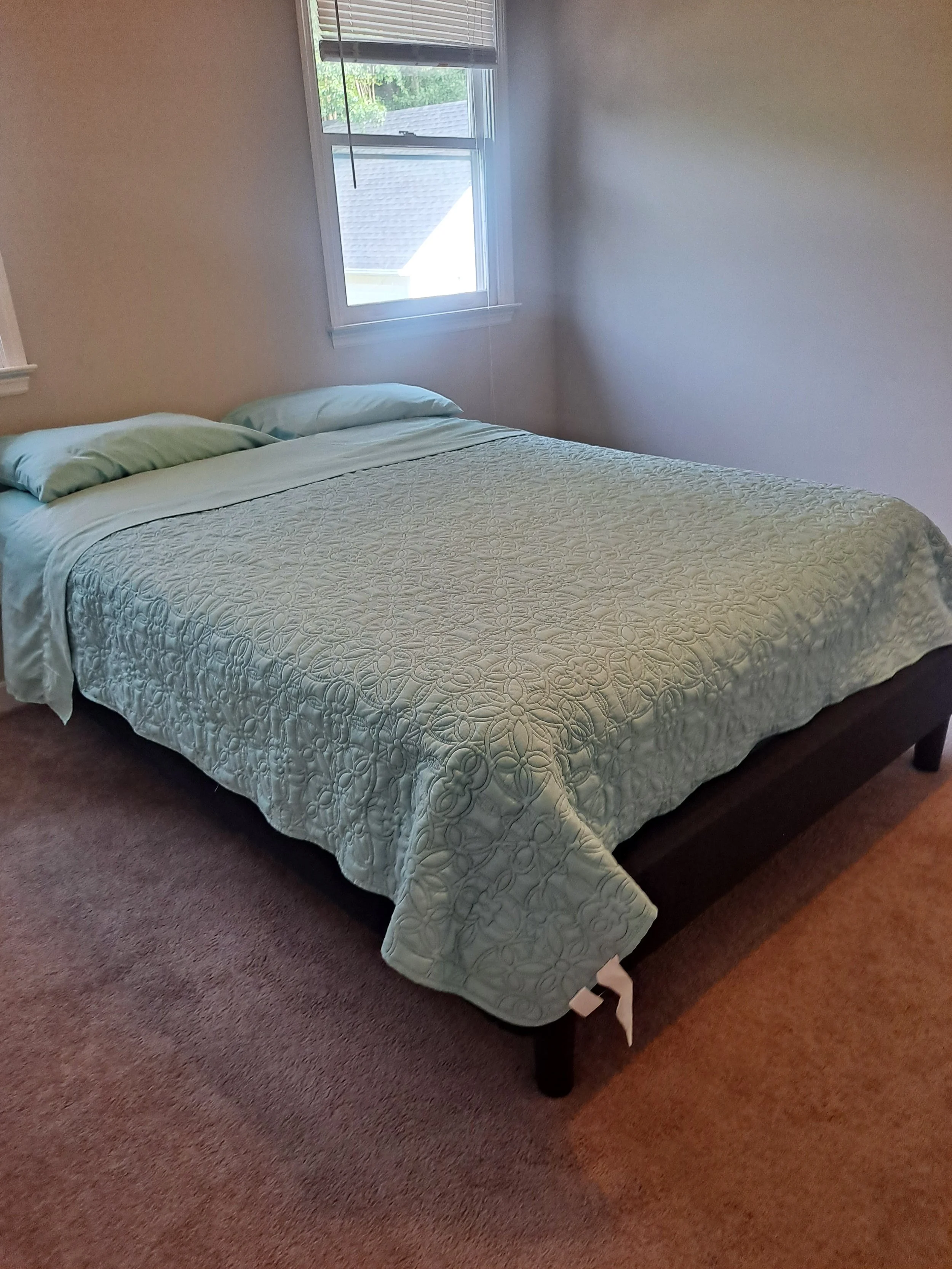 A bedroom with a bed covered with a quilt and two pillows, and a window with blinds letting in natural light.