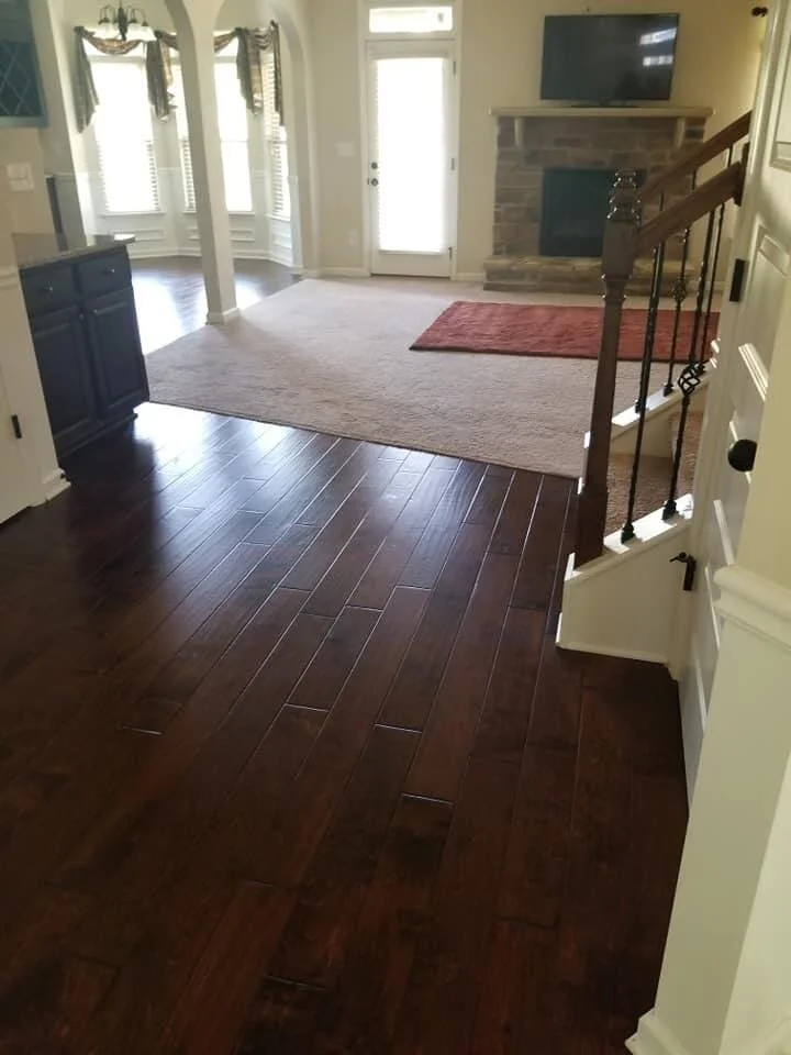 View of a living room with dark hardwood floors, a beige carpet, a stone fireplace with a wall-mounted TV, and a staircase with wooden railing.