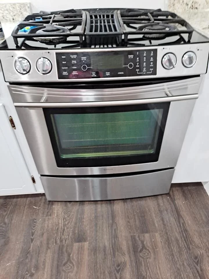 Stainless steel gas stove with four burners on top and a large oven below, positioned between white cabinets on a wood floor.
