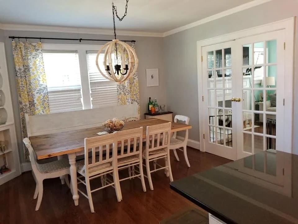 Dining room with a wooden table and eight chairs, two windows with yellow and gray patterned curtains, a chandelier, French doors, and a side table with bottles and decor.