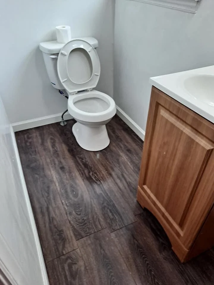A small bathroom with a toilet, a wooden bathroom vanity, and a white sink. Toilet paper on top of the toilet tank. The flooring is dark wood.