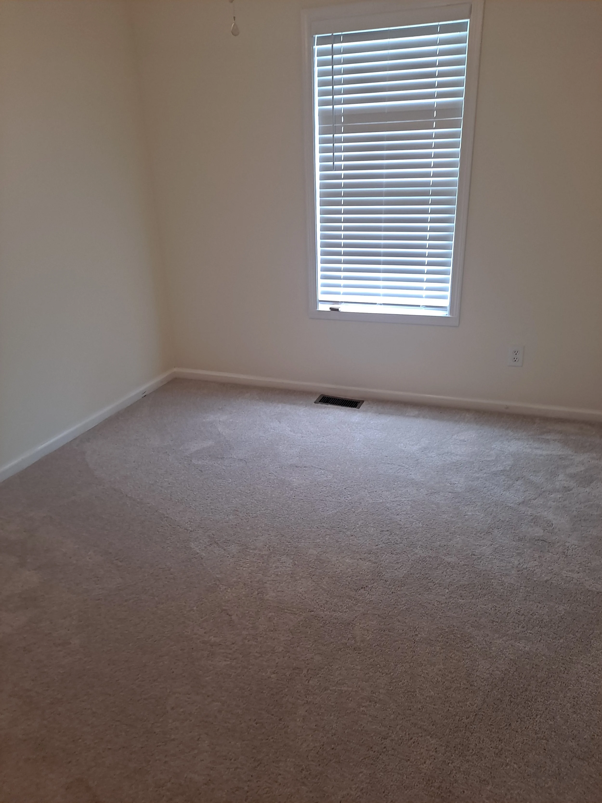 Empty room with beige carpet, white walls, a window with closed blinds, a floor vent, and an electrical outlet.