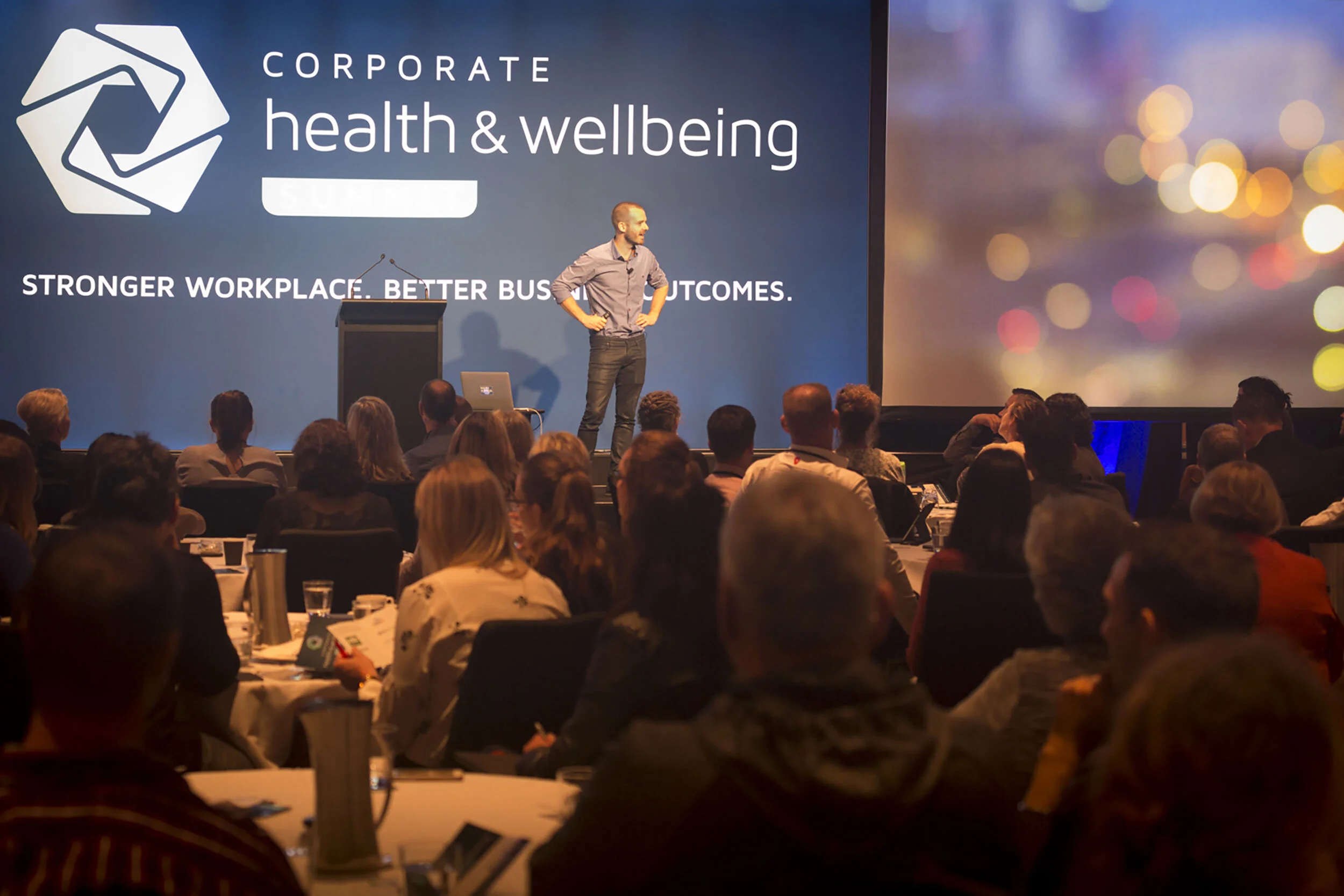 A speaker stands on stage in front of a large screen reading 'Corporate health & wellbeing' during a conference. The audience sits at round tables, listening and taking notes.