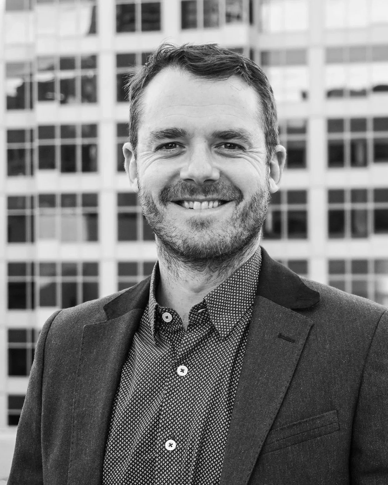 A smiling man in a suit jacket and patterned shirt posing outdoors in front of a modern glass building.