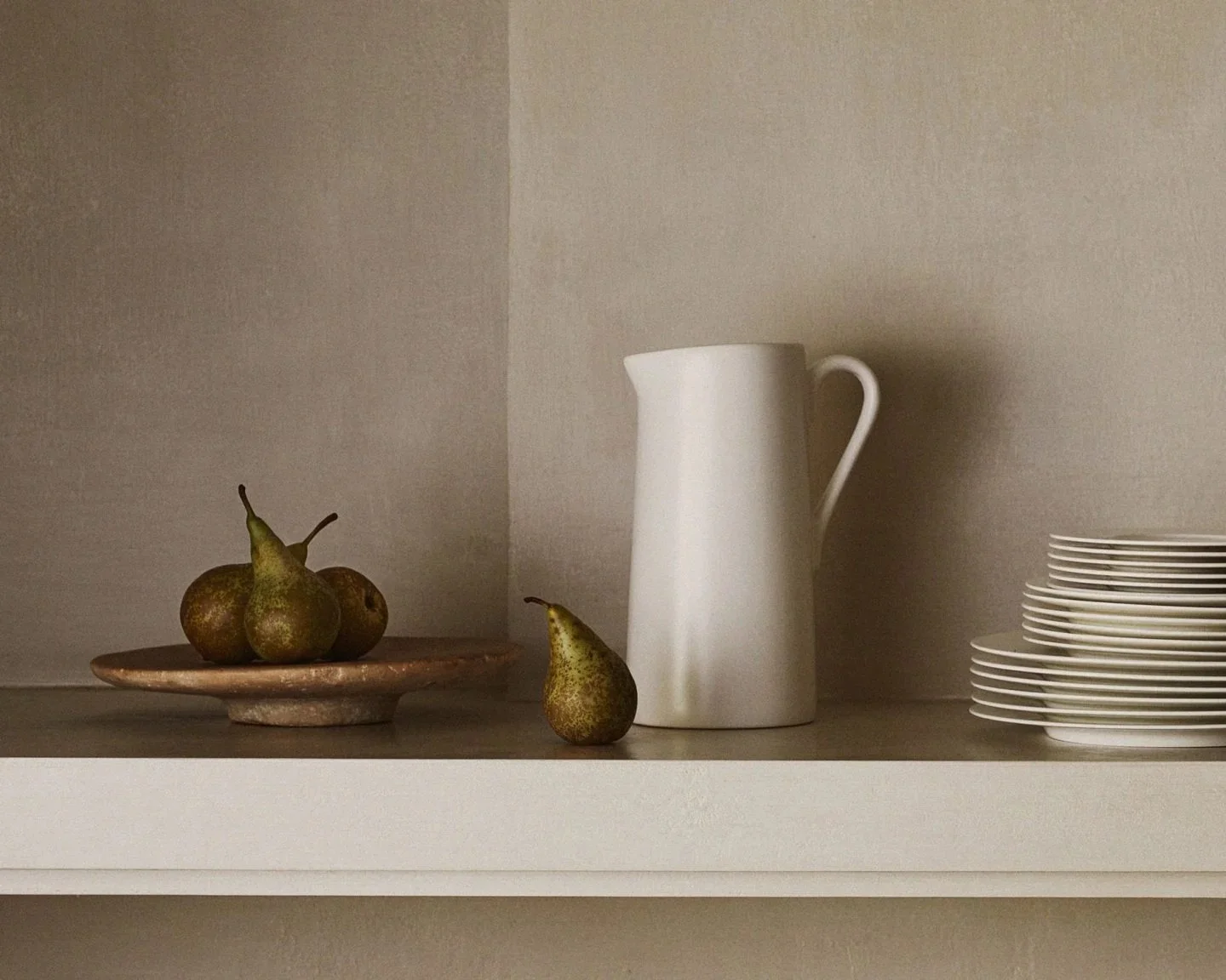 Shelf with three pears on a ceramic plate, one pear outside the plate, a tall white pitcher, and a stack of white plates.