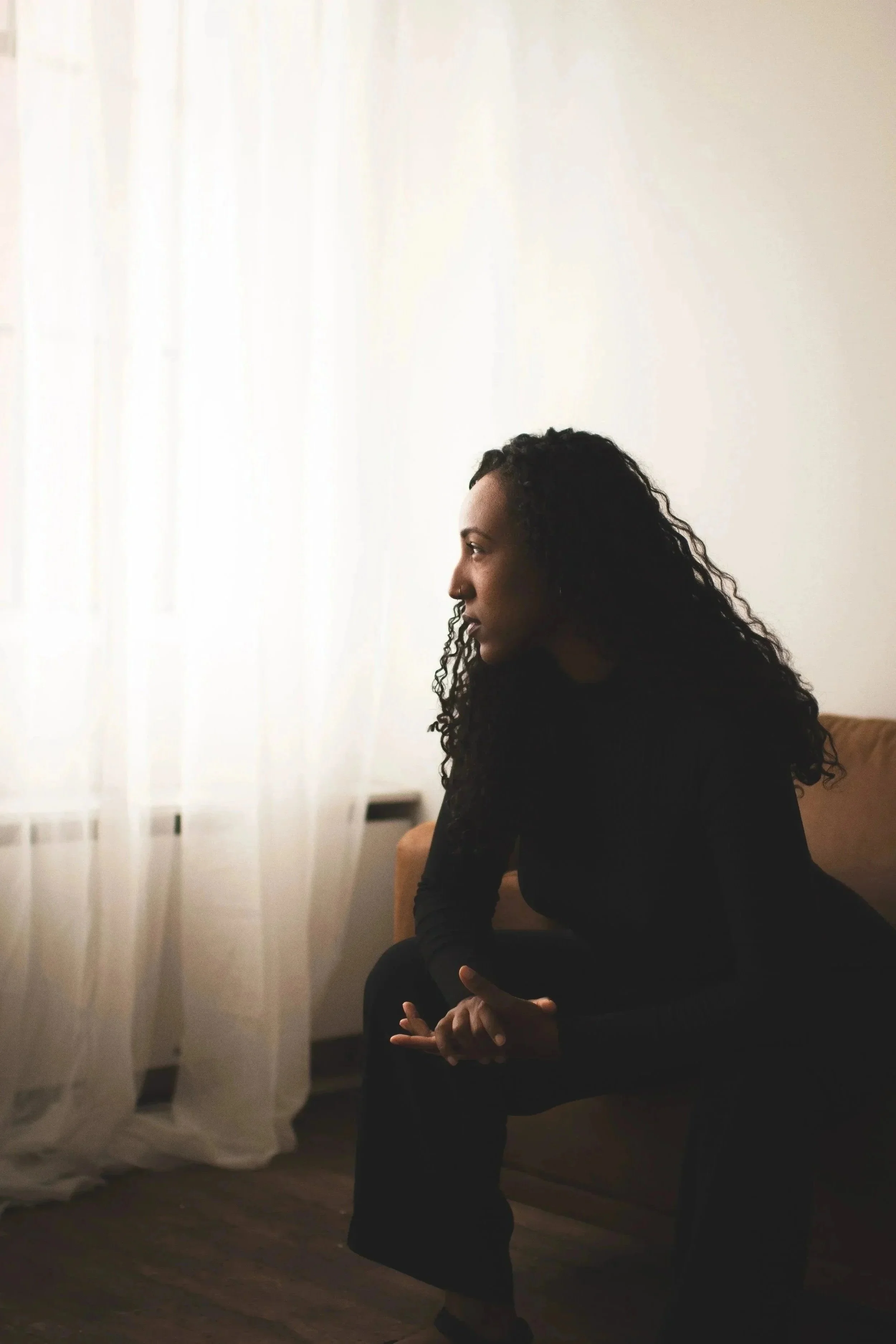 A woman with curly hair sitting with her hands clasped, looking to the left, in a room with white curtains and wooden flooring. House of Nicoline - HON - intuitive guidance, astrocartography, viberise - Jetta Nicoline Harrison