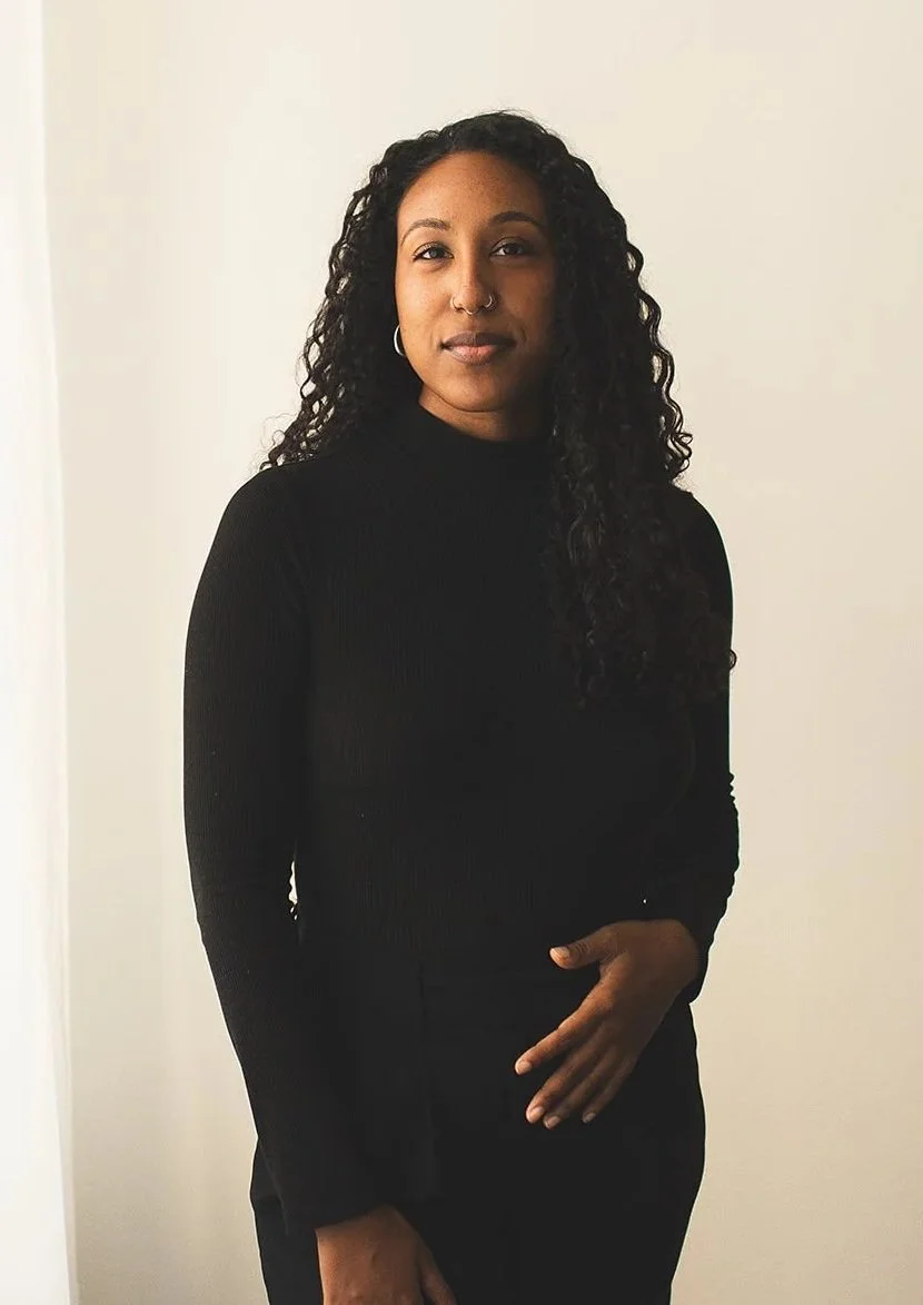 A young Black woman with curly black hair, wearing a black long-sleeve top, standing against a plain off-white wall. House of Nicoline - HON - intuitive guidance, astrocartography, viberise - Jetta Nicoline Harrison