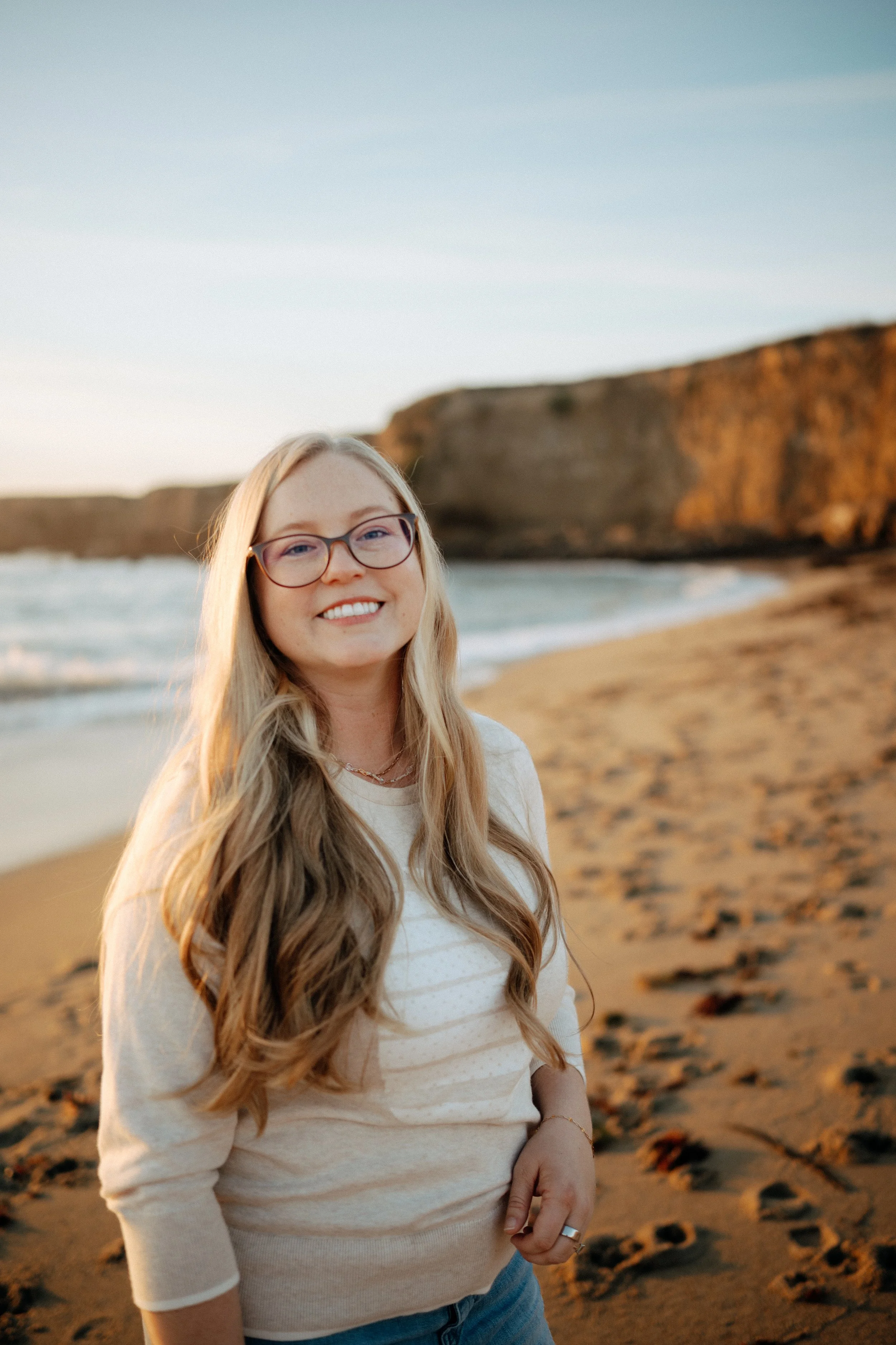 A woman with long blonde hair and glasses smiling on a beach with cliffs in the background during sunset.
