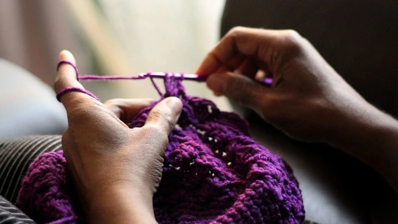 Close-up of person knitting a purple garment, sitting on a striped surface near a window with light filtering through.