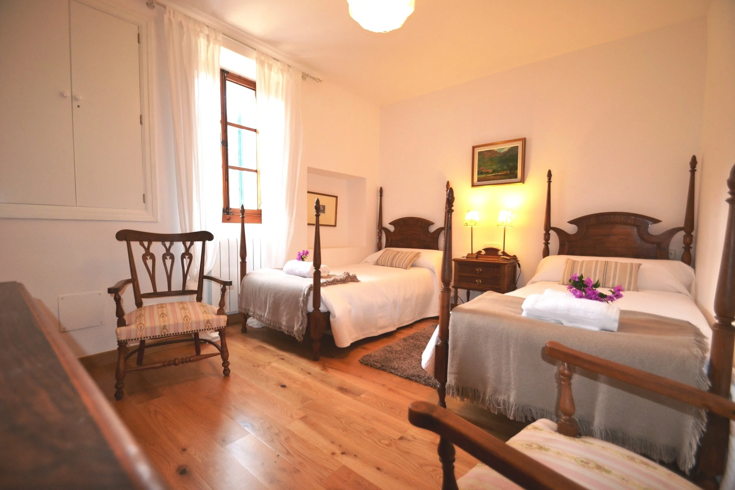 Gemütliches Schlafzimmer mit zwei Einzelbetten, Holzböden, Sessel und Nachttisch. Fenster mit Vorhängen und Wandbilder.