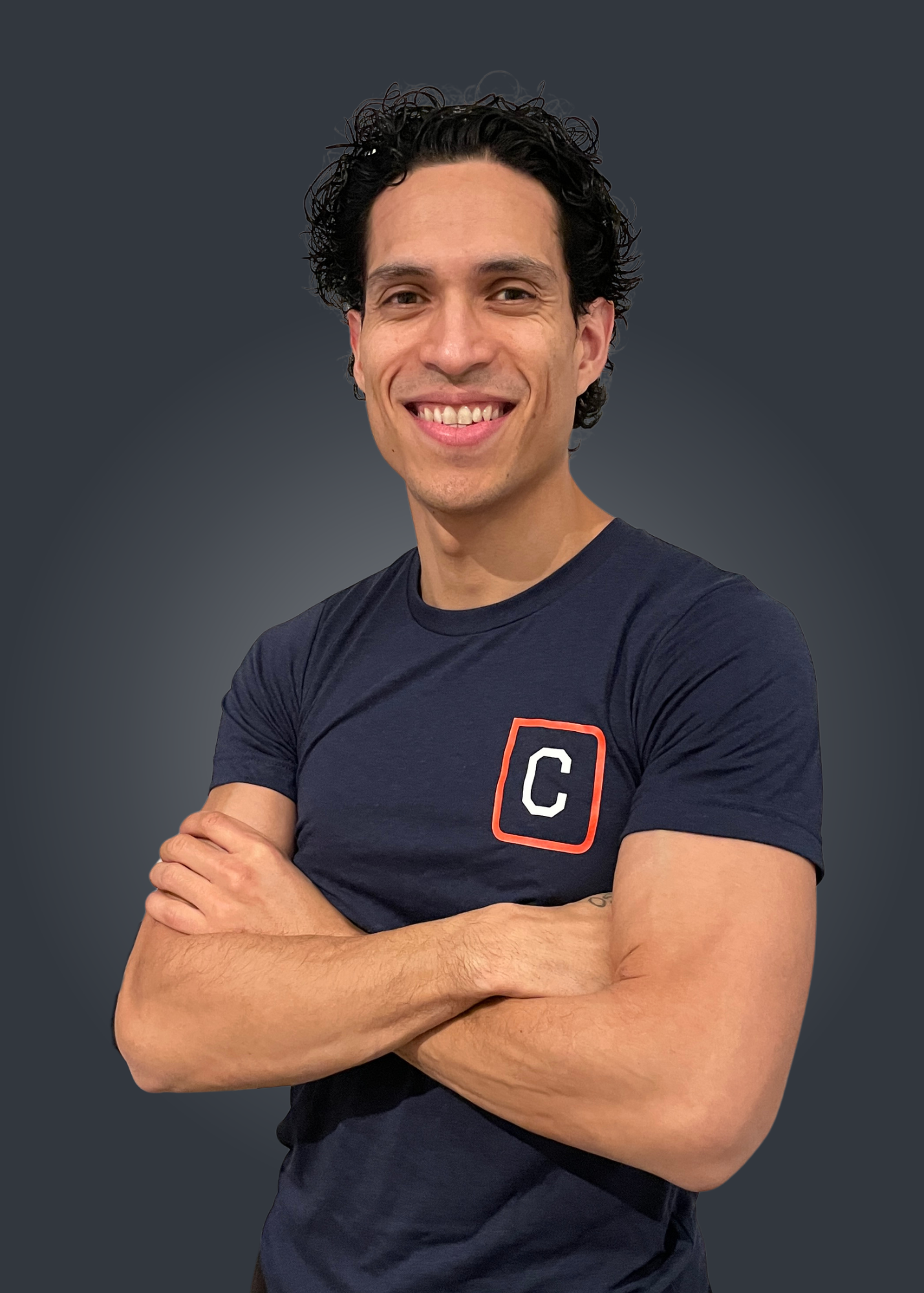 A man with dark curly hair smiling with arms crossed, wearing a navy blue t-shirt with a red and white 'C' logo on the chest, standing against a dark grey background.