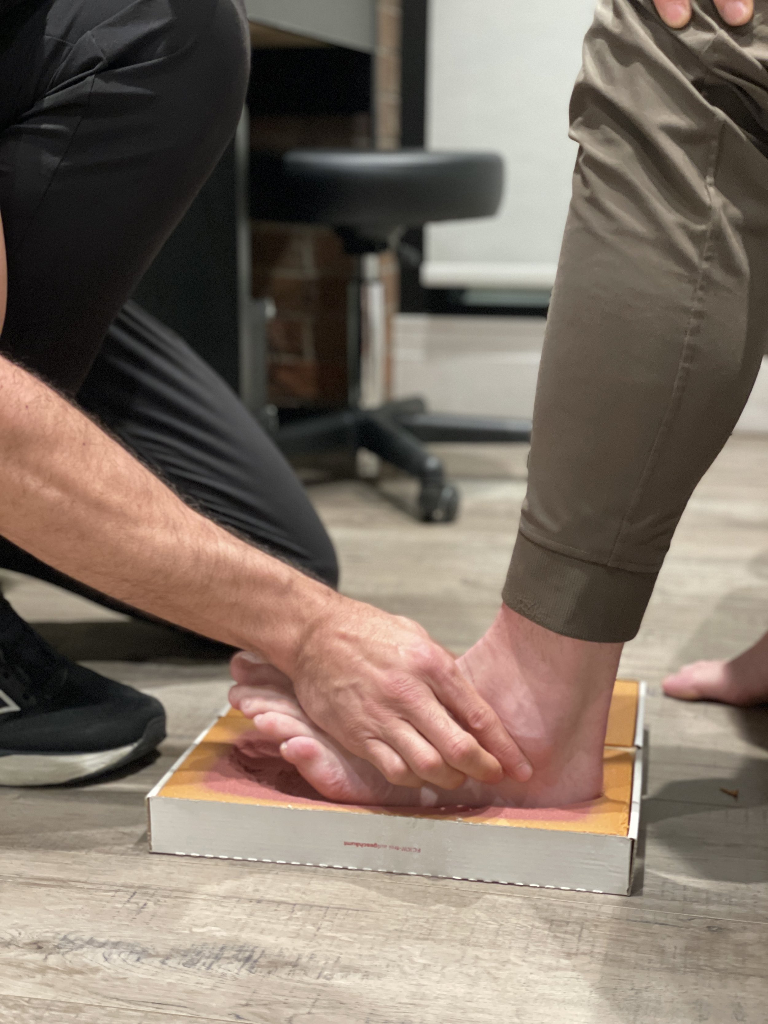 A person comforts another person's bare foot, which is placed on a pizza box on the floor, in an indoor setting.