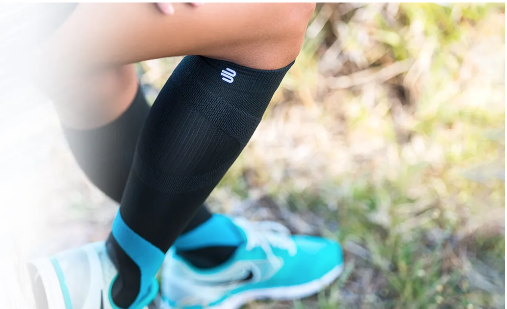 Close-up of a person's lower leg and foot wearing a black compression sock with a small logo, blue running shoes, and standing on a trail with grass and plants.