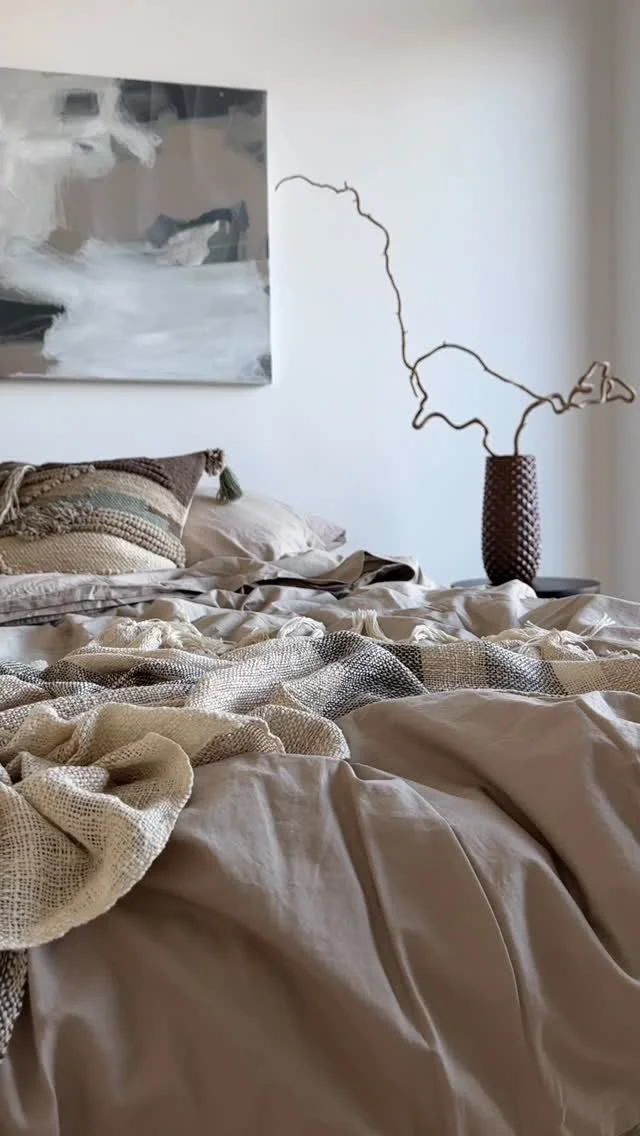 Säng med dekkiga lakan, ett prytt tyg och en kudde, väggdekoration av en tunn, krokig kvist i en krukväxt, och ett abstrakt konstverk på väggen.