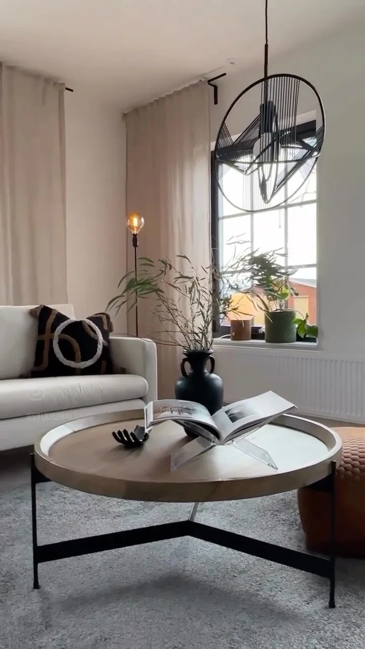 Vardagsrum med vita väggar och stora fönster, ett soffbord med öppna böcker, en svart vas med växter, ett vitt soffbord och en ljus golvlampa, samt ett modernt takpendel.