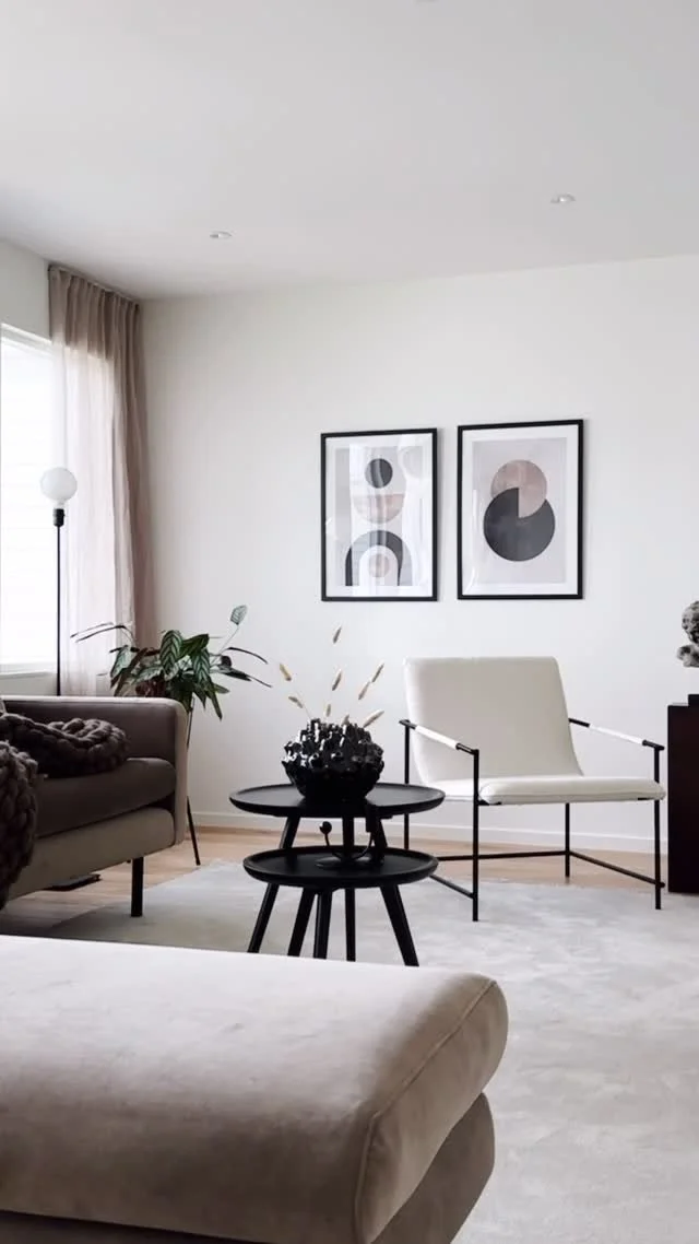 Vardagsrum med vita väggar, två konstnärliga svartvita posters på väggen, en soffa, en stol, ett svart sidebord med en prydnad, ett växter på ett bord, gardiner vid fönstret, en golvlampa.