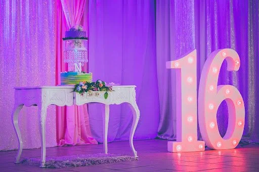 A large lit up 16 decoration is to the right of a small ornate table with the Sweet 16 birthday cake on top.