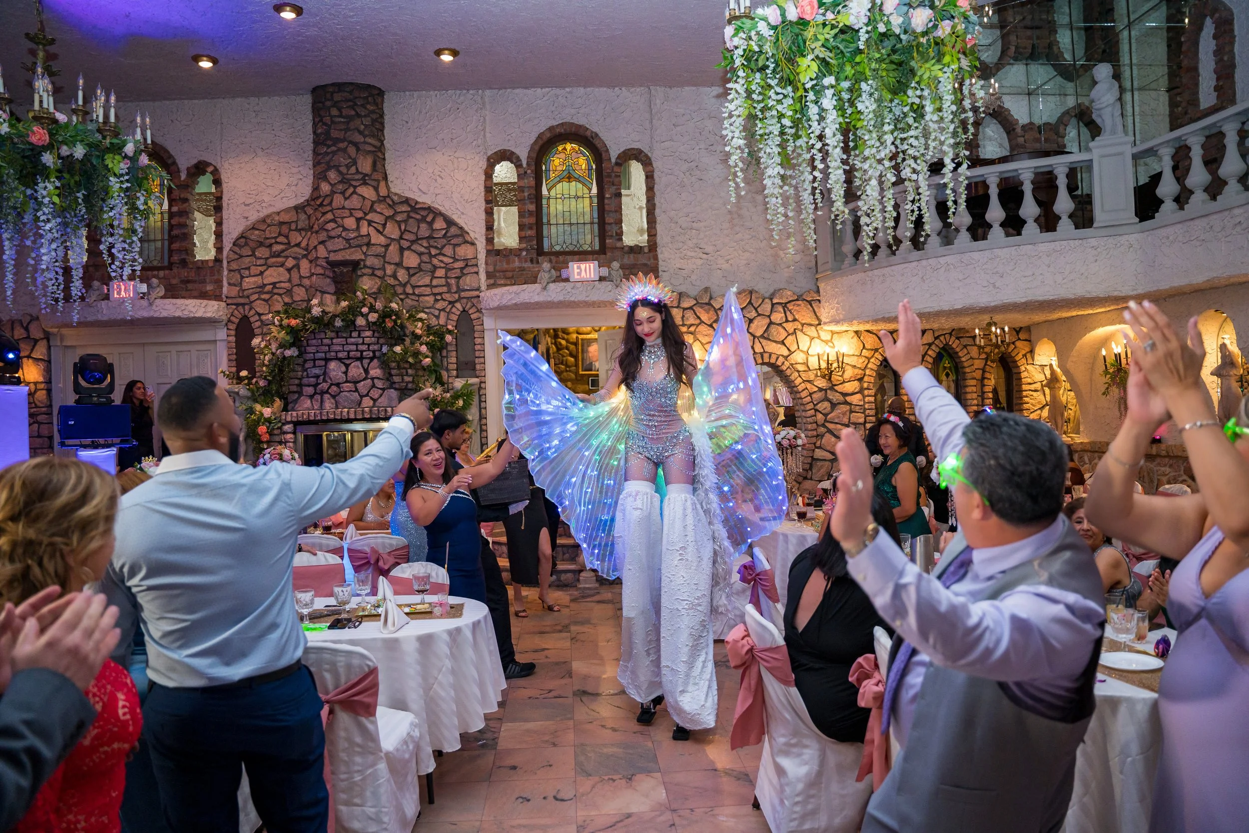 A performer dressed as a fairy with luminous wings and a crown entertains guests at a decorated indoor event with floral arrangements, chandeliers, and stained glass windows in Villa Russo event venue, in Queens NY
