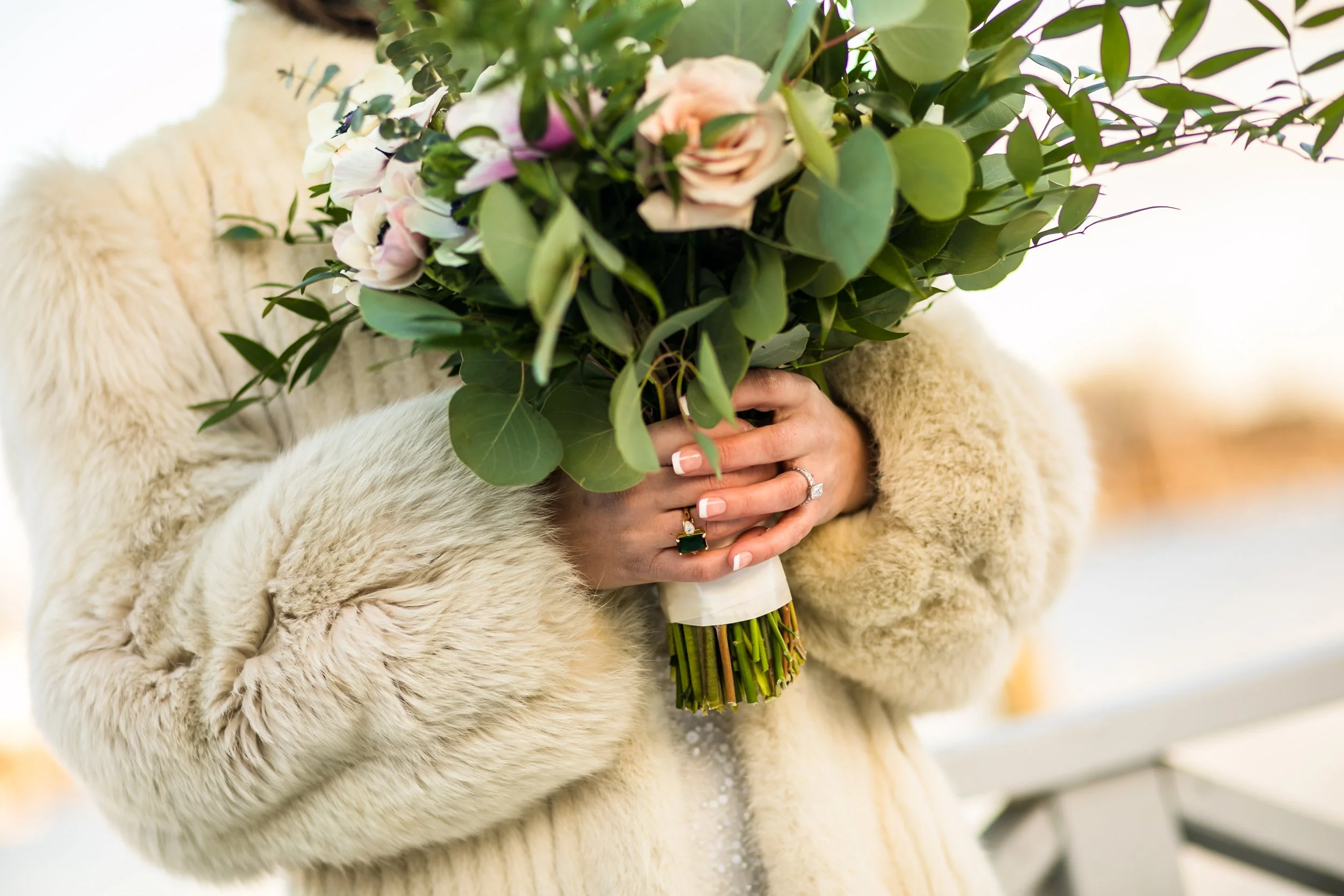 A woman in a fur coat holding a small arrangement of flowers. Her wedding ring is visible.