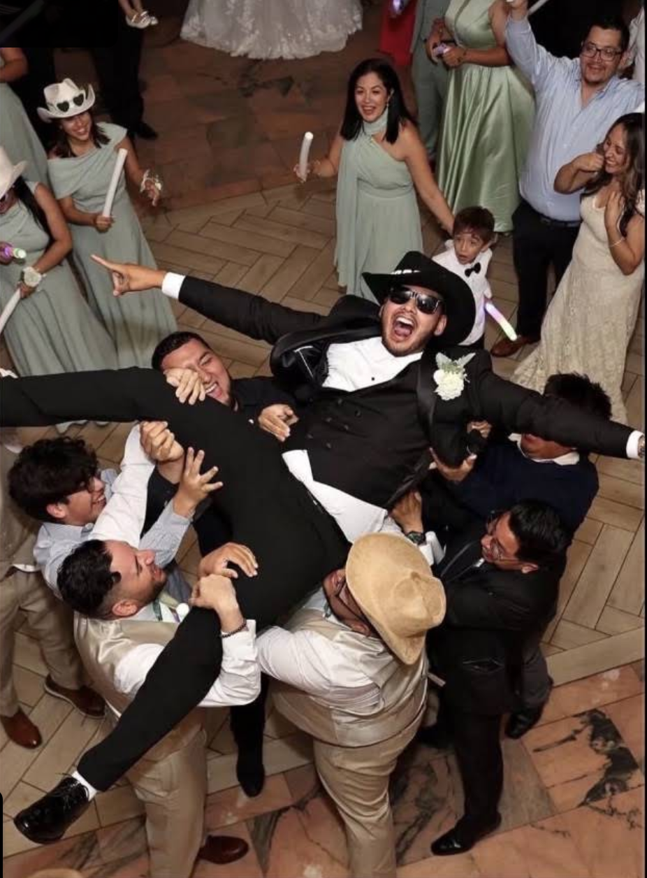 Group of people celebrating at a wedding reception, lifting the groom in a joyous manner, with women in pastel dresses, children holding glow sticks, and guests dressed in formal attire.