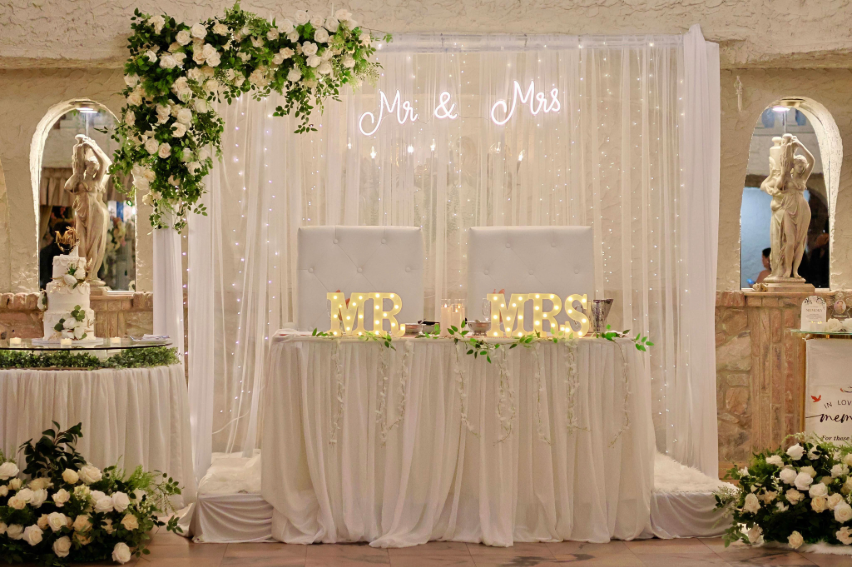 Wedding sweetheart table decorated with white florals, greenery, draping, and illuminated Mr. and Mrs. signs at Villa Russo.