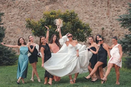 A bride holding a nontraditional bouquet of flowers is surrounded by her bridesmaids, all in different color dresses posing for the photograph.
