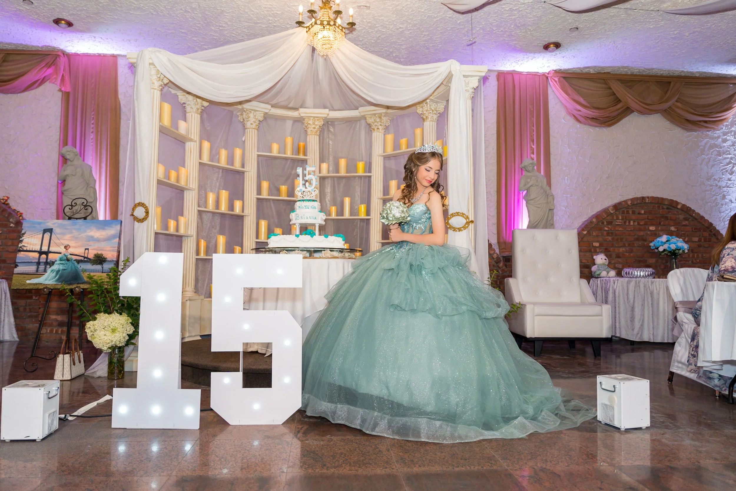 A girl in a teal dress posing in front of her Quinceañera cake and decorations, including a large lit up "15".