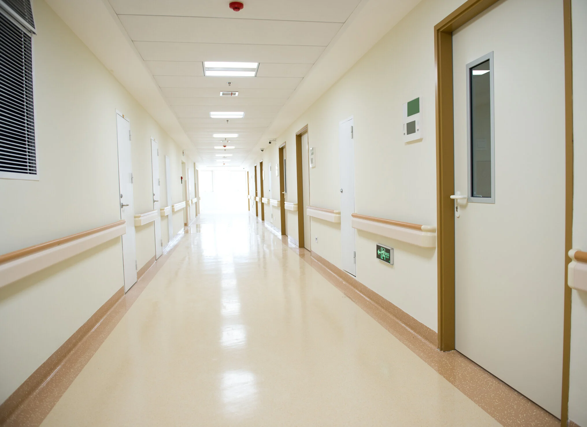 Empty hospital corridor with beige walls, floor, handrails, and multiple closed doors, some with small observation windows and emergency signs, illuminated by ceiling lights.